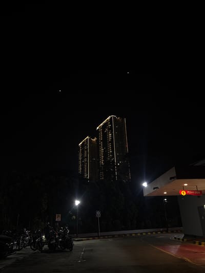 The image depicts a nighttime scene in Bencongan Indah, Indonesia. Two tall buildings with illuminated windows and rooftop lighting are visible in the middle ground. A street light illuminates parked motorcycles. The background includes dark vegetation and a dark sky. A building with a "PESAN DISINI" sign is in the foreground next to a parking area.