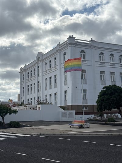 A vertical, medium-shot image shows a multi-story, white building in Arrecife, Spain, likely a government or public building, given its architecture. A rainbow flag hangs from a flagpole attached to the building's facade. The sky is overcast with thick, grey clouds.

To the right of the building, along a sidewalk, a park bench is painted in rainbow colors, adding to the theme of LGBTQ+ pride. Next to the sidewalk is a paved road with white lane markings. A few trees with green foliage are visible, as well as a second flag in the far background that is not discernible.