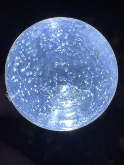 This media file shows a close-up, macro photograph of a transparent, spherical object, possibly a glass marble or bead, against a dark, blurred background. The object is illuminated from behind, revealing numerous small air bubbles trapped within its material. Some of these bubbles are round, while others appear to be elongated or irregular. The surface of the sphere is not perfectly smooth; there are visible scratches and some larger, cloudier inclusions or areas of imperfection within the material itself. In the upper left quadrant of the sphere, there is a small, irregularly shaped yellowish spot. The lighting causes prismatic effects, with faint rainbow-colored refractions visible around some of the bubbles and along the edge of the sphere. At the bottom edge of the sphere, near the right side, a very small dark object, possibly an insect or a piece of debris, is attached or resting on the surface. The overall impression is of a detailed study of a small, man-made object, likely under magnification, with a focus on its internal structure and surface characteristics. There is no visible text, and the setting provides no specific context for time of day, weather, or location beyond the provided context of Branipole, Bulgaria, which is not visually indicated in the image.