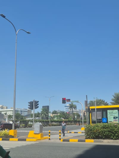 The image depicts an urban street scene in Pasay, Philippines on a clear, sunny day. A man in a patterned shirt and dark pants is crossing a pedestrian crosswalk. Traffic lights are visible, with the closest set showing a red light. There is a bus stop with a yellow roof structure along the street, featuring advertisements and signage. A sign reads "MACAPAGAL BLVD." The scene includes palm trees, streetlights, various buildings in the background, and yellow and black painted road dividers. A bus stop is also visible.