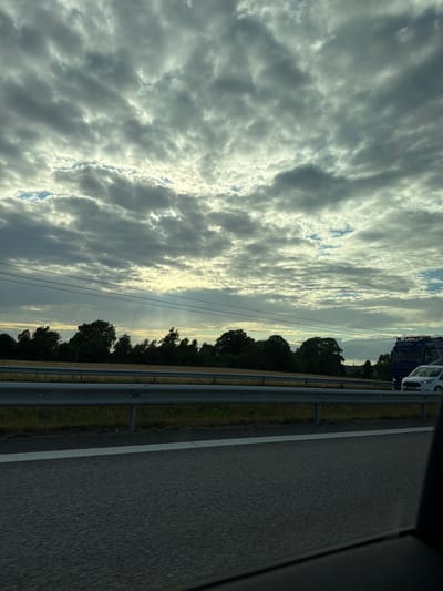 The image captures a view from a vehicle traveling on a highway, likely near Himle, Sweden, given the context provided. The sky is predominantly filled with heavy, gray clouds, with breaks allowing shafts of sunlight to pierce through, illuminating portions of the horizon and creating a dramatic, atmospheric effect. Below the sky, a line of trees forms a dark silhouette against the lighter sky. A metal guardrail separates the highway from the grassy embankment and fields beyond. In the distance, on the highway, a blue truck with a white cab is visible, along with a white vehicle in front of it. The road surface is asphalt with a white lane marker. The perspective suggests the photo was taken while in motion.