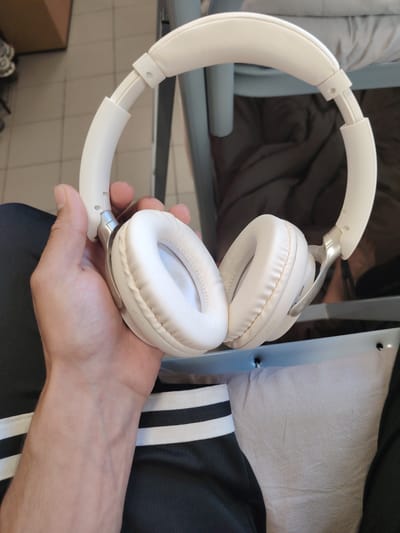 A person's hand holds a pair of white over-ear headphones with a silver accent. The headphones are positioned in the center of the frame, with the headband arching over a light-colored surface. The person's arm is visible in the lower left, wearing black pants with white stripes. The background is blurred, but appears to be an indoor setting with furniture. The location in the city of Часцы, Russia is not determinable from the image.