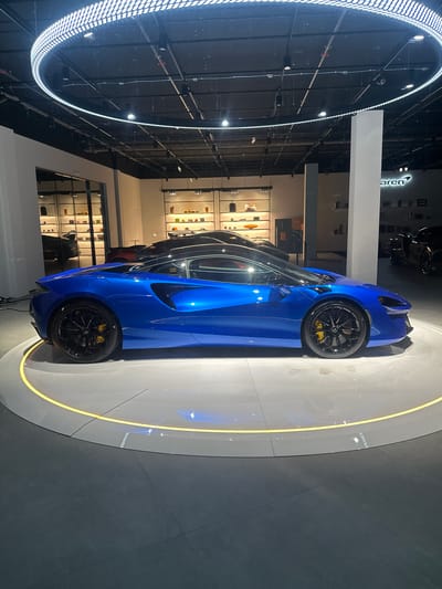 The image shows a blue McLaren sports car on a circular platform with a yellow outline. The car is positioned at an angle relative to the camera. The location appears to be a McLaren dealership, as the brand logo is visible in the background. Additionally, there are display shelves with car accessories. The building structure features a circular light fixture on the ceiling. Based on the location information, the scene likely takes place in Glattbrugg, Switzerland.