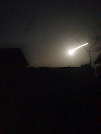 An eerie, nighttime scene features a faint, angled streak of light piercing through fog or mist. The light originates near tree branches silhouetted in the upper portion of the frame. A dark, shadowy structure or building dominates the right side of the image, sharply contrasting with the illuminated left side. The overall impression is mysterious and otherworldly.