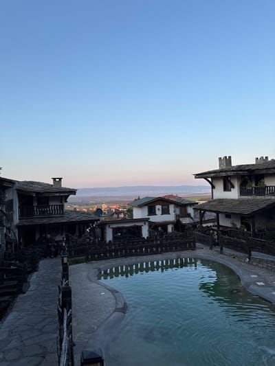 A vertically-oriented, angled outdoor shot depicts a building complex in Medovo, Bulgaria, with a stone-edged pool of water at the right. The scene is illuminated by the soft, ambient light of dusk or dawn, casting a subtle gradient of color across the sky.
The architecture consists of multi-storied, possibly traditional buildings with dark, stone-like shingle roofs and white walls. Dark wooden balconies and fences line the facade facing towards the pool. The pool itself is filled with opaque light-blue water, reflecting the ambient light.
In the background, a distant landscape with light green and brown hues suggests a valley or plain, bordered by faint mountains and a horizon where the sky meets the land. No people are clearly discernible within the frame, but a gathering of indistinct forms in dark clothes can be seen under an overhang. The environment is a blend of constructed elements and natural scenery.