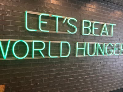 The image displays a vibrant neon sign affixed to a dark, textured brick wall. The wall is composed of dark grey or black bricks, possibly painted, arranged in a horizontal pattern. The neon sign consists of two lines of text, rendered in a glowing green or aqua color. Both lines of text are presented in an upside-down orientation as seen in the photograph.
The top line of text reads "WORLD HUNGER," and the bottom line reads "LET'S BEAT." The letters are formed by neon tubes mounted on a clear or white backing, which is then attached to the brick wall. The bright luminescence of the sign contrasts sharply with the dark background, creating an eye-catching visual. There are no people or specific activities taking place; the image focuses solely on the illuminated signage.
The setting appears to be an interior or exterior wall of a building, likely a commercial establishment or public space, designed to convey a message. The time of day is not explicitly clear, but the glowing sign suggests it is either dark or dusk, or the sign is simply designed to be illuminated constantly. This scene is located in Kelvin Grove, Australia.