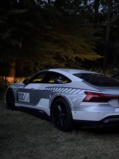 An Audi e-tron GT electric vehicle, partially covered in a grey and white camouflage wrap with purple accents, is parked on a grassy area at night. The car is viewed from the rear three-quarter angle, with its sleek design and distinctive taillights illuminated. The background consists of dark trees and foliage, suggesting an outdoor setting. The scene is illuminated by artificial light, likely from a nearby source, casting a warm glow on the car and the surrounding environment. The "e-tron" badging is visible on the lower side of the vehicle. The car appears to be in good condition, with no visible damage. The location is identified as Mensdorf, Luxembourg.