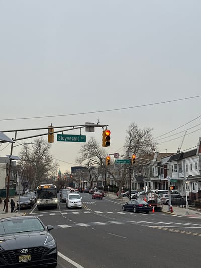An urban street scene in Jersey City, United States, depicts a busy intersection under an overcast sky. The image captures a street with multiple lanes, filled with cars and a bus, all appearing to be stopped at a red traffic light. The time of day appears to be daytime, but the muted lighting suggests either an overcast day or perhaps early morning/late afternoon.

In the foreground, a black SUV with a New Jersey license plate "B99RLX" is visible on the right. Directly behind it, a white SUV and a public transit bus are seen. The bus displays "87 Journal Sq" on its destination sign. Other vehicles are lined up further down the street, indicating moderate traffic congestion.

Prominently featured are multiple traffic lights; the set directly in front shows a red light illuminated, while a smaller set on the cross-street shows an amber or yellow light. A green street sign hanging over the intersection reads "Stuyvesant Ave." Another street sign in the distance also indicates "Stuyvesant Ave" and "57th St." A "No Left Turn" symbol is also visible near one of the distant traffic lights.

The street is lined with multi-story buildings, likely a mix of residential and commercial, with classic urban architecture. Bare-branched trees are scattered along the sidewalks, suggesting late fall or winter. In the background, a construction crane can be seen above the rooftops. The scene conveys a typical urban environment with daily traffic.