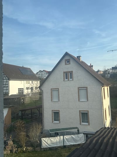This image depicts a residential scene in Krauchenwies, Germany, viewed from an elevated perspective under a clear blue sky with faint cirrus clouds and contrails.
In the immediate foreground, the ridged dark gray roof of a building, likely the observer's location, is visible in the lower right. Adjacent to it, a patch of green grass with sparse, leafless deciduous shrubs and rockery is present. A low fence, constructed from a dark wire mesh covered with a white fabric or tarp, spans horizontally across the lower midground. Behind this fence, a green-roofed lean-to structure stands at the base of the prominent building.
The central subject is a two-story, light-colored (off-white or cream stucco) residential building with a steep, dark brown gabled roof. The front facade features several rectangular windows with brown-trimmed surrounds: one small square window at the apex of the gable, two vertically stacked rectangular windows below it, and two larger rectangular windows on the second floor, both fitted with rolled-down external white shutters. On the first floor, two more large rectangular windows are visible, each revealing internal curtains. A dark downspout runs vertically along the right side of the building. Direct sunlight illuminates the left side of this building, casting clear shadows.
In the background, to the left of the central building, stands a white residential house with partial dark timber framing on its left facade and a dark gabled roof equipped with multiple solar panels. Further behind these structures, a gently sloping hillside is populated with additional residential buildings featuring predominantly brown or reddish-brown tiled roofs. A tall, white and red/orange construction crane is visible against the sky on the far right horizon, indicating ongoing development in the distance. No individuals, actions, or direct interactions are visible within the frame.