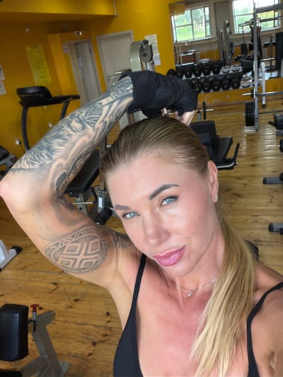 In a gym, a Caucasian woman with blonde hair pulled back in a ponytail and blue eyes poses for a selfie. She is wearing a black sports bra and a black wrist wrap on her left arm, which is extended upwards, revealing extensive tribal tattoos. Her right arm is out of frame. The gym features yellow walls, wooden floors, and various weightlifting equipment, including dumbbells, weight racks, and cardio machines visible in the background. The setting is likely a small, local gym.