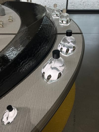 The scene is an interior display area located in Seongdong-gu, South Korea. The primary subject is a large, curved display counter featuring a central, irregularly textured, reflective black surface that resembles rippled water. This black section is bordered by a lighter gray, horizontally ribbed surface. Six clear glass bottles are positioned on the ribbed surface, including one partially visible in the lower left. Each bottle contains a translucent liquid and is designed with a distinctive base incorporating three metallic, pointed claws. The bottles have two types of caps: three are textured black, and two (further back) are light-colored. Several small, light-colored, amorphous objects are visible on the far side of the central black display section. The background reveals a dark gray floor with a partial curved yellow marking, and a large window allowing ambient light into the space. No individuals or active interactions are present.