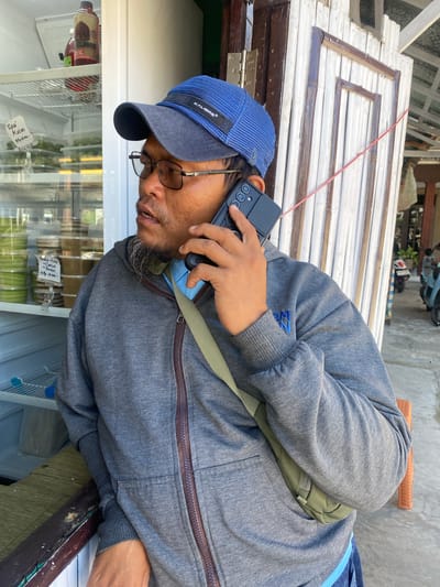 A man with a beard and glasses is standing and holding a cell phone to his ear, seemingly in conversation. He is wearing a blue baseball cap and a grey hooded sweatshirt. A green bag is slung across his chest. In the background, there is a white wooden door, a refrigerator filled with food containers, and a glimpse of motorbikes parked outside. The setting appears to be a street-side stall or shop. The image is taken outdoors in what appears to be a tropical climate, given the presence of a fan and bright sunlight.