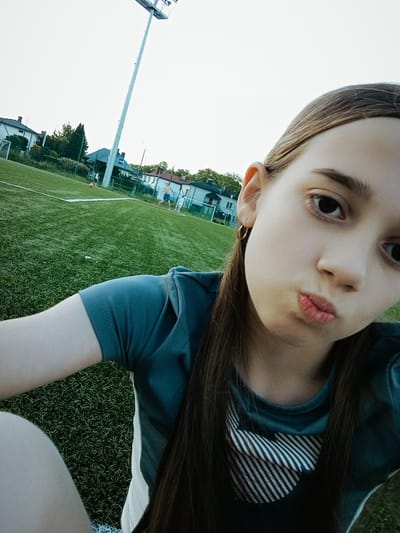 The media file is a selfie taken by a young person on a grassy field. The subject is lying down, with their upper body and head in the foreground. They are wearing a teal t-shirt with a white striped heart design on the chest. Their hair is long and dark brown, falling across their shoulders. The person is looking directly at the camera with their lips pursed and their eyes open. A subtle golden hoop earring is visible in their right ear.
In the background, the field appears to be a sports field, possibly a soccer field, with white boundary lines visible. Further in the distance, there are buildings and trees, suggesting a suburban or semi-rural environment. A tall light pole with a lighting fixture is also prominent in the background. The sky is bright and overcast, indicating daytime. The overall impression is that of a casual, outdoor setting. The photo has a slightly distorted perspective, typical of a selfie taken at a wide angle.