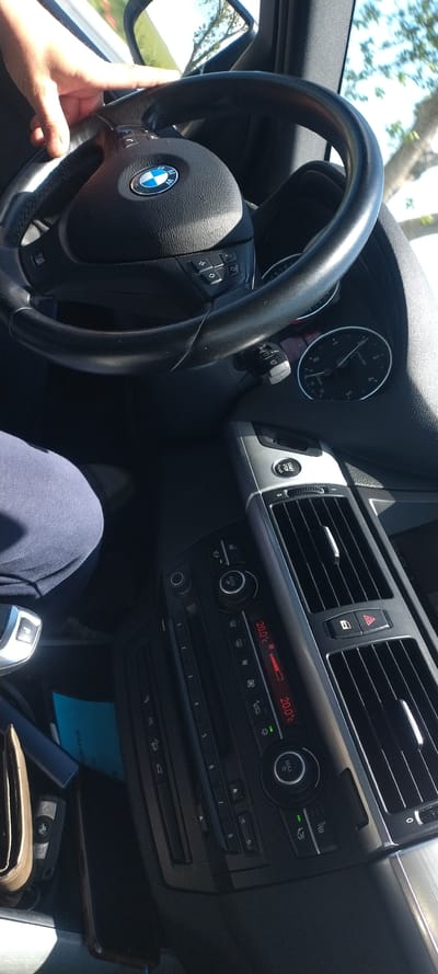 The image captures the interior of a BMW car. The driver's hands are visible on the steering wheel, which bears the BMW logo. The dashboard features a speedometer, a start/stop button, air vents, and climate control settings, displaying a temperature of 20.0°C. Below the climate control, there's a radio panel and storage space containing a phone, wallet, keys, and a blue card. The gearshift is in "P" for Park. The photo's location in the city of Neofit Rilski, Bulgaria, is not discernible from the visual elements within the image.