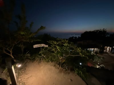 The image shows a twilight view over the sea in Karimunjawa, Indonesia. In the foreground, there is a dirt path illuminated by a spotlight. To the left, a tree with sparse foliage is visible. A signpost with "LOR Lunge Dret Ratopo" written on it stands near a cluster of green plants. In the background, silhouettes of trees and buildings are seen against the dusky sky. The sea is calm with a few lights visible on the water, possibly from boats. The overall scene is peaceful and serene.