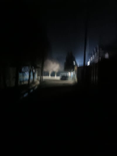 A blurry night scene depicts a dark street. Light illuminates the road ahead, revealing trees and distant structures. Buildings line the right side of the street with illuminated windows. Shadows dominate the foreground, obscuring details on the left. The overall impression is of a quiet, slightly eerie, nighttime environment.