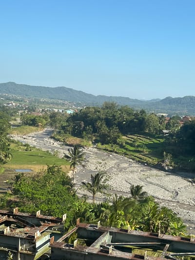 The image depicts a landscape scene, likely captured from an elevated vantage point, featuring a riverbed with dry rocky sections and some flowing water, surrounded by lush green vegetation and rice paddies. In the background, a town or village is nestled at the base of rolling, tree-covered hills under a clear blue sky. The foreground shows the rusty, weathered framework of a bridge or industrial structure. The scene is devoid of any people or animals. The location is identified as Adisana, Indonesia.