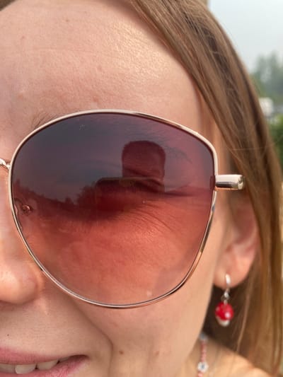 A close-up, head-and-shoulders shot focuses on a person wearing large, rose-tinted aviator sunglasses. The sunglasses reflect a blurry landscape, possibly trees and sky. The person's skin, visible on the forehead, cheek, and upper lip, shows texture and a few faint freckles. A single eyelash is visible above the sunglass lens. To the right of the sunglasses, the person's ear is visible with a dangling earring featuring a small, red and white mushroom charm. The person's hair is brown and appears slightly wavy, falling around their face and shoulders. A hint of a pink and white beaded necklace is visible below the chin. The background is blurred, suggesting an outdoor setting with a soft, overcast sky. The image is cropped such that only a portion of the face and hair is visible. There are no discernible landmarks indicating the city of Tartar, Russia.