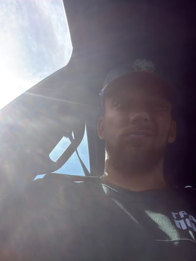 An over-the-shoulder, selfie-style, medium shot depicts a man wearing a dark baseball cap with a crest on it, and a dark t-shirt with white lettering. The man is looking upwards and to his left, with his face partially obscured by bright sunlight streaming in from the upper left of the frame. The background is dark, suggesting the interior of a vehicle, with a bright sky visible through a window. The lettering on the t-shirt appears to be "ГРА ПОР". The scene is lit by strong sunlight, creating lens flare and overexposure in parts of the image. The location is the village of Varvara, Bulgaria, within what appears to be a vehicle.
