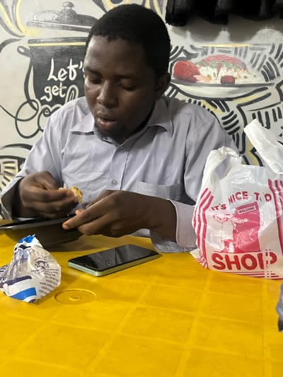An indoor portrait from Barako, Nigeria, features a young man with dark skin, dressed in a light grey long-sleeved collared shirt. He is seated at a textured yellow table, seemingly focused on eating or preparing a snack. He holds a small, light-colored food item in his right hand, while his left hand grasps a dark object, possibly a utensil or another piece of food.
On the yellow table, a smartphone with a light green/teal case lies face down, alongside a crumpled blue and white snack wrapper. Visible text on the wrapper includes "NIGERIA," "BIDA," "GOVERNMENT MINNA," and "ASTATE NIGERIA," confirming the regional context. A white plastic shopping bag with red text, featuring phrases like "SHOP" and "IT'S NICE TO WELCOME," rests near the man's arm.
The background wall is adorned with abstract black graphic patterns and some colorful elements. Notable wall art includes an illustration of a food dish and the hand-lettered phrase "Let's get." The lighting suggests an indoor setting, likely under artificial light. The man appears calm and engrossed in his activity.