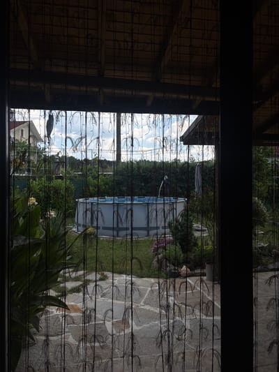The image depicts a backyard scene viewed through a beaded curtain, suggesting an outdoor living space or covered patio. In the foreground, the beaded curtain is a prominent feature, obscuring parts of the view and creating a textured overlay. Beyond the curtain, a partially visible house with a light-colored facade and a red-tiled roof is situated on the left. The middle ground features a large, circular above-ground swimming pool with a blue-patterned liner. Surrounding the pool is lush greenery, including manicured hedges, shrubs, and a stone-paved pathway. Various plants, some with large leaves and others with flowers, are also visible. The sky above is partly cloudy and bright, indicating daytime. The overall environment suggests a suburban or rural residential setting, likely in Viden, Bulgaria, given the context. There are no people or animals present in the image, and no activities are being performed.