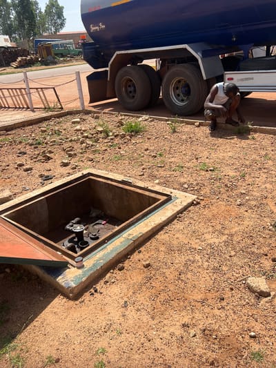 The image depicts a scene in Mbar, Nigeria, featuring a large blue tank truck parked near a ground-level, open, rectangular concrete enclosure. A man wearing a white tank top and shorts is kneeling near the truck's wheels, appearing to be working on something. The open enclosure contains pipes and other mechanical components, potentially related to a utility or fuel system. The ground is covered in reddish-brown dirt and gravel. In the background, there are additional vehicles and stacked firewood along a paved road.