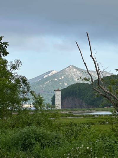 The image shows a serene landscape with a body of water in the foreground, likely the Avacha Bay, with lush green vegetation and trees on the banks. In the middle ground, a stark, white, rectangular structure, possibly a former industrial building or observation tower, stands near the water. Behind it, a steep, tree-covered hill rises, and beyond that, a large, snow-dusted mountain dominates the background, characteristic of the volcanic landscape around Vilyuchinsk, Russia, a city known for its proximity to active volcanoes. The sky is overcast with a soft, diffused light, suggesting a cloudy or misty day. The scene evokes a sense of natural beauty and a touch of industrial history.