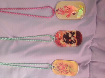 The image displays three dog tag-style pendants hanging from beaded necklaces. The top pendant features a cartoonish pink bird with large eyes and wide wings against a pale yellow background with iridescent highlights. The middle pendant shows a black and white cartoon character with spiky hair and fierce eyes surrounded by red and yellow abstract shapes. The bottom pendant has a yellow background with a cartoon character in shades of pink and red, appearing to be in action with dynamic lines. All pendants are oval with rounded corners and have a hole at the top for the necklace attachment. The necklaces are made of small, round beads; the top one is pink, and the other two are light green. The pendants and necklaces are laid out on a textured, light purple fabric surface, likely a blanket or cloth, which appears to be draped or folded, creating soft shadows. The image is taken from a top-down perspective, and there are no visible indications of the city of Inhulo-Kamianka, Ukraine.