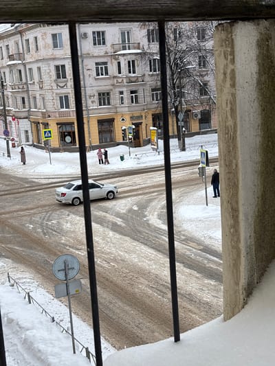 An elevated, downward-angled view captures a snow-covered intersection in Velikiye Luki, Russia, likely the junction of Prospekt Lenina and Oktyabr'skaya Ulitsa, centered on the building at 19 Prospekt Lenina. The foreground is partially obscured by three black vertical metal railings and a concrete pillar on the right, with accumulated snow on the immediate lower edge.
The scene depicts a winter day, with snow covering all sidewalks, verges, and rooftops. The road surfaces at the intersection are a mix of dirty slush, exposed pavement, and visible tire tracks, indicating vehicular traffic. A multi-story, light-colored corner building with classical architectural elements and signs of exterior wear dominates the background. Its ground floor, painted ochre, hosts businesses, including one marked "ПИВНИЦА" (Brewery/Beer House).
Four individuals are present:
1. Two persons, one in a pink coat and one in a darker coat, stand together on the snow-covered sidewalk near the "ПИВНИЦА" entrance, appearing to interact.
2. A solitary person in a long, light-brown coat stands on the far left sidewalk, near a pedestrian crossing sign.
3. A male individual in a dark jacket and blue jeans stands on the right sidewalk, positioned near a pole with traffic signals.
A white sedan, consistent with a Lada Granta, is mid-traverse across the intersection, moving from right to left. Multiple street signs are visible, including several yellow diamond-shaped pedestrian crossing markers, a red "No Entry" sign (partially visible), and a circular sign with a cross-like symbol in the lower-center foreground. A green trash receptacle is positioned near the two individuals at the corner, and a yellow advertising stand is also present there. Bare deciduous trees are visible behind the buildings.