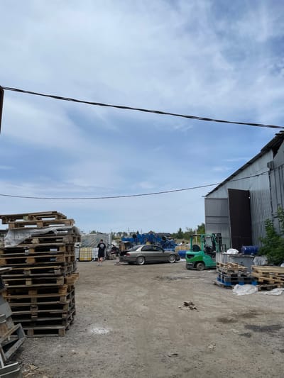 The image depicts an outdoor industrial scene, likely a storage or logistics yard, in Кенен Әзірбаев ауылы, Kazakhstan. A tall stack of wooden pallets is prominently positioned on the left side of the frame. In the middle ground, a person in a black t-shirt and shorts stands near a light-colored sedan with its trunk open. A green forklift is parked to the right of the sedan, facing the viewer. Adjacent to the forklift, more stacks of wooden pallets are visible. In the background, a large, corrugated metal industrial building dominates the right side of the image. Beyond the building and to the left, numerous large blue barrels are stacked in rows. The ground is composed of dirt and gravel. The sky is partly cloudy.