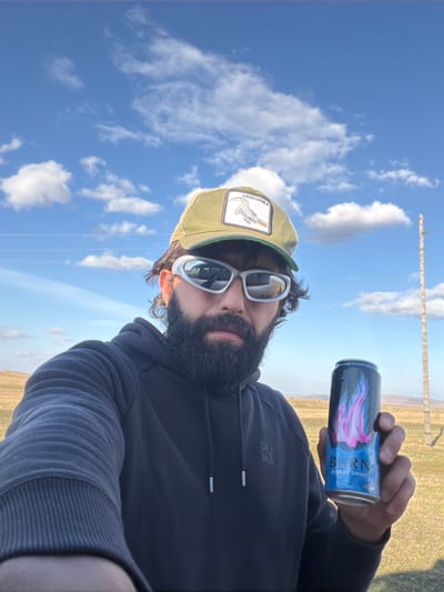 A selfie photo depicts a bearded man with sunglasses and a Crocodile-branded hat holding a "BURN" energy drink. The background reveals a landscape with a blue sky, white clouds, a grassy field, and a utility pole. It's daytime. There's no specific indication this scene is taking place in the city of Знаменка, Russia. The man wears a dark Puma hoodie.
