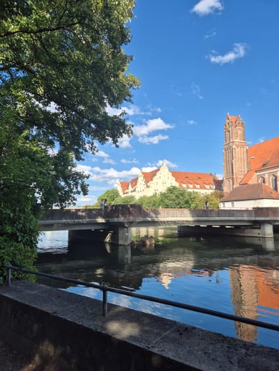 The image depicts a scene in Landshut, Germany, featuring a bridge crossing a body of water. On the right side of the frame, a tall brick tower with a red tiled roof rises prominently, adjacent to a building with a similar red tiled roof. To the left of the tower, a large, pale yellow building with multiple red gabled roofs and numerous windows is visible, nestled behind lush green trees. A concrete bridge spans the water, supported by pillars. The water reflects the sky, clouds, and the buildings and bridge above. In the foreground, a stone embankment with a black metal railing lines the water's edge. A large, leafy tree occupies the upper left portion of the image. Two individuals can be seen standing on the bridge, facing away from the camera, and another individual is walking on the bridge. The sky is bright blue with scattered white clouds.