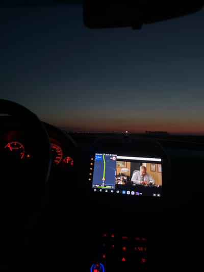 The media file depicts the interior of a car at dusk. The scene is dominated by the car's dashboard and a central infotainment screen. On the infotainment screen, a video is playing featuring two people. One person, wearing a light-colored collared shirt, is seated at a table and appears to be engaged in a conversation or presentation. Behind them, framed pictures hang on the wall. Below the video, a navigation map is displayed, showing a route with turn-by-turn directions. The time on the screen reads "22:06".

The dashboard instruments, illuminated in red, include a speedometer and a tachometer. The speedometer needle is pointing towards the lower end of its range, indicating a low speed. The ambient light outside the car suggests it is either dawn or dusk, with a gradient of colors in the sky from dark blue to orange near the horizon. A line of distant lights stretches across the landscape.

The setting appears to be a road in or near Побалаўскі сельскі Савет, Belarus, given the location context. The overall mood is one of quiet travel during the evening. The presence of the infotainment screen with both entertainment and navigation suggests the occupants are on a journey. No visible emotions are discernible from the subjects in the video due to the distance and resolution.