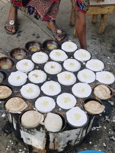An overhead shot captures a vendor cooking small, round, white pancakes with a golden center, using a specialized pan with multiple hemispherical molds. Some pancakes are still being cooked, while others are already golden brown. The vendor's hands and feet, adorned with sandals and colorful clothing, are visible on the sides. A weathered concrete surface and a wooden stool are also in view, suggesting an outdoor setting.