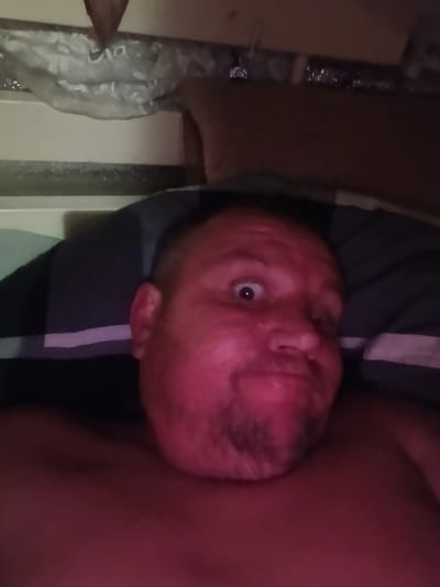 A close-up, low-angle selfie taken from bed shows a man's face, which is illuminated with a bright pinkish-red light. The man is lying on a pillow with a dark pillowcase that has a white stripe. He has light-colored skin, brown hair, and a beard stubble. His eyes are wide open, and his mouth is slightly pursed. The background is dark and blurry, with hints of bedding or furniture. The location in the city of Vilne, Ukraine is not determinable from the image.