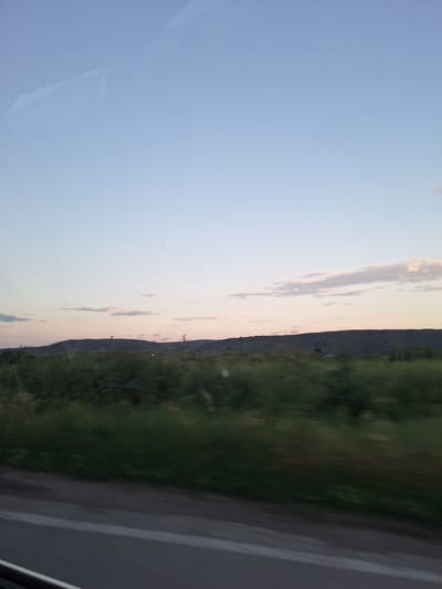An outdoor landscape image, captured from inside a moving vehicle at dusk, depicts a rural or semi-rural scene in the vicinity of Topoli, Bulgaria. The sky, occupying the upper two-fifths of the frame, transitions from clear light blue at the zenith to pale pink and orange hues along the horizon, with scattered, elongated clouds exhibiting soft white and pink tones. Faint rectangular reflections are visible on the window surface in the upper left sky.

A prominent line of dark green, densely vegetated hills or low mountains spans the entire width of the midground, forming the horizon. Two tall, slender metallic structures, likely electrical pylons, are positioned on the hills: one in the left-center and another towards the right-center. A small cluster of distant buildings is faintly discernible on the right side, against the backdrop of the hills.

The foreground is characterized by a band of dark green vegetation, likely bushes or trees, exhibiting significant horizontal motion blur, indicating the speed of the vantage point. Below this vegetation, a dark grey asphalt road occupies the bottom portion of the image, featuring a faint white line towards the right edge. A dark, indeterminate structure, likely part of the vehicle's interior, is partially visible at the bottom left corner. No individuals or animals are visible in the scene.