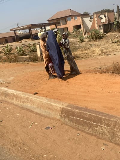 This daytime image, taken in Lapai, Nigeria, depicts an outdoor scene featuring three individuals on a reddish-brown dirt ground.
In the foreground, a worn, light-colored concrete curb runs horizontally, separating a lower dirt area from the main dirt expanse where the individuals stand. Scattered small debris is visible on the ground.
In the mid-ground, three individuals, likely adult females, are gathered. The individual on the left wears a light brown tunic over dark trousers and has a light-colored, possibly white, bundle balanced on their head. They face towards the center. The central individual is largely obscured by a dark blue, full-length garment. The individual on the right wears a patterned, full-length dress and head covering, facing the central figure with hands raised, appearing to be speaking or gesturing. They all cast distinct shadows to their right, indicating strong sunlight from the left.
The background reveals several structures. To the left, a large brown sign structure with the text "MA'AJIYA GLOBAL COMPANY LIMITED" is visible, marking what appears to be an entrance or gate. Behind this sign, a dark cylindrical tank and a single-story building are partially visible. To the right of the sign, a two-story building with a reddish-brown tiled roof and light brown walls stands. Further to the right, a partially destroyed or unfinished building with exposed rebar and damaged walls is present. Sparse green vegetation grows along the roadside and near the background buildings. The sky is clear.