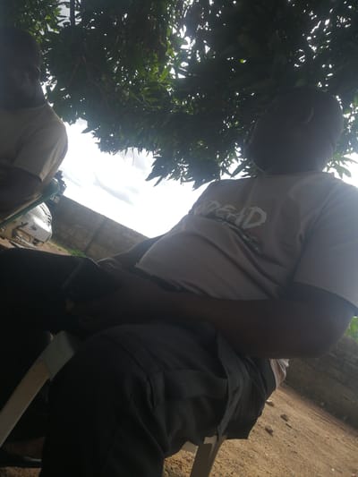 Two individuals are seated outdoors under the shade of leafy trees. One person, in the foreground and to the right, is wearing a light-colored t-shirt with visible graphic text that appears to read "DEAD" in distressed white lettering. This person is also wearing dark pants and is holding a dark object, possibly a phone, in their lap. Their head is turned to the right, and they appear to be looking upwards or off to the side. Another person is partially visible on the left, wearing a light-colored t-shirt. Behind the individuals, a white vehicle with visible damage is parked near a brick wall. The ground appears to be dry and sandy, with scattered pebbles. The sky above is cloudy and bright, suggesting it is daytime. The presence of the trees and the open, dry ground suggest a rural or semi-rural setting in Ajasse Ipo, Nigeria. The overall atmosphere seems relaxed, with the subjects seeking shade from the sun.