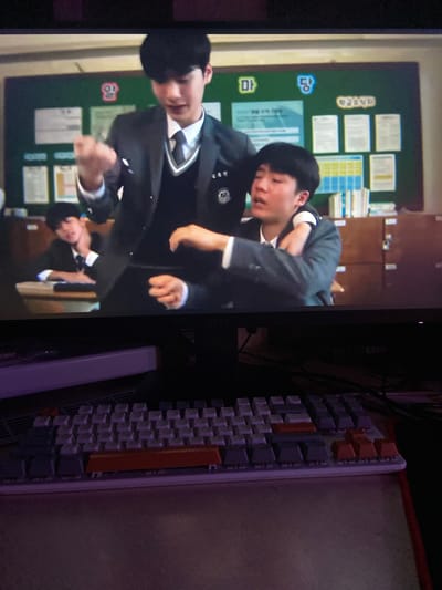 In the foreground, a portion of a white and purple mechanical keyboard is visible, with several keys illuminated with an orange tint. The background shows a television screen displaying a scene from a Korean drama. Two young males in school uniforms are depicted in a classroom setting. One is standing and gesturing with his right hand, while the other is seated, with his arm around the standing male's shoulder. The classroom has a green chalkboard with posters and papers displayed on it, and wooden cabinets filled with books. The scene is consistent with a school environment in South Korea, not Beloretsk, Russia.