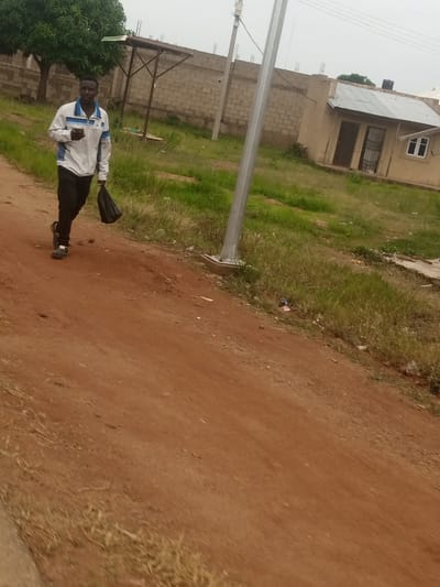 A person, appearing to be a young man, walks on a dirt road in Russo, Nigeria. He is wearing a white jacket with blue stripes on the collar and sleeves, dark pants, and dark shoes. He is carrying a black plastic bag in his right hand. The road is made of reddish-brown dirt, with dry grass and some sparse vegetation on the sides. In the background, there is a wall constructed from concrete blocks, a small building with a corrugated metal roof, and a tall metal pole, likely a streetlight or utility pole. A large green tree is visible to the left of the scene. The sky is overcast.