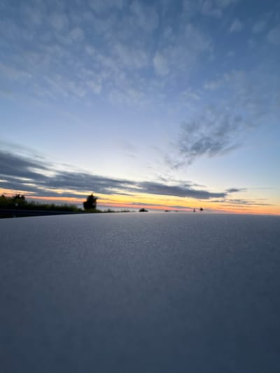 The image is a low-angle, close-up shot of a frosted surface, likely the hood of a car, with a blurred background of a sunrise over the Baltic Sea in Paldiski, Estonia. The foreground is dominated by the textured, gray frosted surface, which fills the lower half of the frame. Beyond this, a strip of land with trees and a dark silhouette of a structure is visible, bordering a band of colorful sky. The horizon line is low, with hues of orange, yellow, and pink radiating from the sunrise, transitioning into shades of blue and gray in the clouds above. The overall atmosphere is serene and cool, indicative of an early morning. No people are visible in the image.