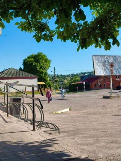 A bright, sunny outdoor scene in Velyka Bahachka, Ukraine, captures a quiet moment in what appears to be a small town or village square. The time of day is likely mid-morning or early afternoon, indicated by the clear blue sky and strong, distinct shadows cast across the paved ground in the foreground.

In the immediate foreground on the left, a modern metal bicycle rack with multiple curved slots is visible on a grey tiled sidewalk. Next to it, a dark trash bin stands. Above, lush green leaves of a large tree, possibly a horse chestnut, frame the top portion of the image, contributing to the shadows on the ground.

Two individuals are seen walking across the open, mostly unpaved area in the mid-ground. On the left, a person dressed in a magenta-colored tracksuit walks away from the viewer, carrying a dark bag or box. To their right, another person in lighter attire (blue and white) also walks away, leading a small, light-colored dog on a leash.

The background features several buildings and a tree-covered hill under the clear sky. To the left, a white building with a horizontally striped green roof and a red band below its eaves is visible. An air conditioning unit is attached to its side. On the right, a large, weathered, rectangular billboard stands on a concrete base, its surface peeling and rusted, making its original content unreadable. Beyond the billboard, a reddish-brown brick building with a dark roof and several windows houses what appears to be a commercial establishment. Partial Cyrillic text, possibly "ПИВНИЙ ЦЕНТР" (Beer Center) or "КОМП'ЮТЕРНИЙ ЦЕНТР" (Computer Center), along with a phone number (e.g., "095 864 58 60"), is faintly visible on a sign on this building. Red circular traffic signs are also present near this building. A dense green tree stands prominent in the center-left background, and utility poles with wires run along the street leading up a gentle incline.