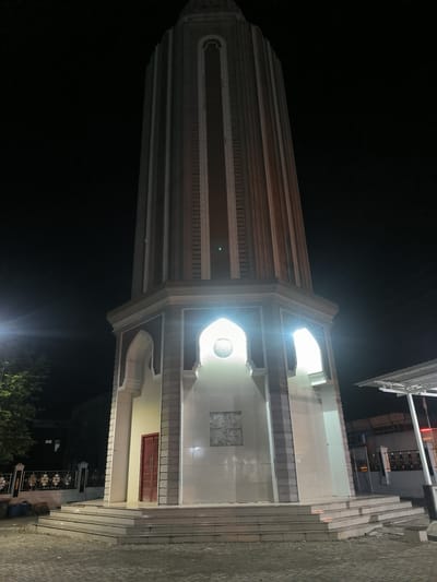 This is a vertical, medium-shot, eye-level, outdoor, nighttime photograph of a tall, octagonal tower with a conical roof. The tower is illuminated by lights from within and around its base. The tower's base is adorned with intricate architectural details, including arched doorways and decorative panels. The tower is surrounded by a paved area and some vegetation. The sky is dark, with a few faint stars visible. The location is specified as "Barat Yaman, Indonesia," which translates to "West Yemen" in Indonesian. However, this likely refers to a specific local place name rather than the country of Yemen. The scene is empty of people.