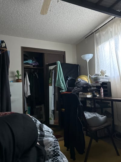 The image depicts a cluttered bedroom. The scene is viewed from a low angle, focusing on a person lying on a bed covered with a black, white, and gray patterned comforter. To the left, a portion of a white dresser is visible with a small potted cactus on top. Adjacent to the dresser is a closet with sliding doors, revealing various clothing items hanging and folded within. On the right side of the image, a desk is covered with numerous items, including papers, containers, headphones, and a pink spray bottle. A floor lamp stands behind the desk, and sheer white curtains hang by a window. The overall impression is one of disarray. The provided context that this scene is located in Vancouver, United States of America is insufficient for a definitive forensic analysis of the location.