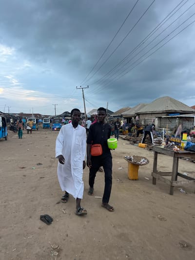 This is a medium shot, eye-level, outdoor photograph taken in the city of Oko Amakom, Nigeria. The scene depicts a dirt road lined with market stalls and buildings. The sky is overcast with heavy clouds. Two young men are walking towards the camera in the foreground. The man on the left is wearing a long white robe and sandals. The man on the right is wearing a black long-sleeved shirt and pants, also with sandals, and is carrying a bright green bucket and an orange bag across his chest. In the background, several blue and yellow motorized tricycles, known as "keke," are parked or moving. There are various market goods displayed on wooden tables and on the ground, including produce like yams, bottles of oil, and other items. Several other individuals are visible in the background, some appearing to be vendors and customers interacting in the marketplace. The overall impression is of a busy, local market day.