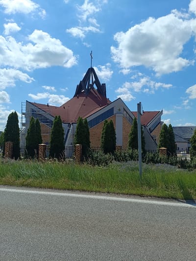 A wide shot captures a modern church under a bright blue sky dotted with fluffy white clouds. The church features a unique architectural style with a multi-peaked, rust-colored roof and light brown brick walls. A prominent cross sits atop the highest peak. Scaffolding is visible on the left side of the building, suggesting ongoing construction or maintenance.
In front of the church, a row of tall, slender evergreen trees stands behind a black metal fence. The fence is supported by brick pillars. Beyond the fence, a strip of green grass and low-lying purple flowers lines the foreground, separating the church property from a paved road. The road, with a white line marking its edge, takes up the bottom portion of the image.
The scene appears to be set in the daytime, with clear weather and ample sunlight. The overall atmosphere is peaceful and serene. No people are clearly visible in the image. There is no visible text. The location is identified as Lipno, Poland.