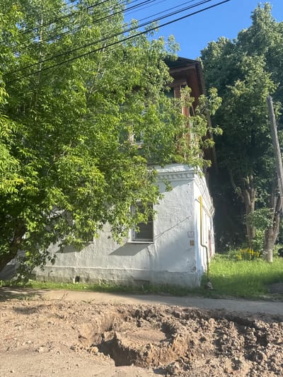 An exterior, eye-level, daytime shot captures the side of a two-story stucco building with a brown wooden mansard roof and dark trim. The building is situated in the city of Vereya, Russia, and appears to be in a residential area. A large deciduous tree with lush green foliage partially obscures the left side of the building. Power lines are visible overhead, intersecting with the tree branches. To the right of the building, a utility pole stands tall, and more trees with dense green canopies create a backdrop. A yellow pipe runs vertically down the side of the building, with a red pipe connected to it lower down. A sign with Cyrillic lettering, reading "ПГ № 16", is affixed to the wall below the yellow pipe. The ground in front of the building is a paved surface, possibly a sidewalk or road, with a section of disturbed earth and a manhole cover visible in the foreground, suggesting ongoing construction or utility work. The overall impression is a somewhat aged, yet lived-in, urban environment bathed in bright sunlight.