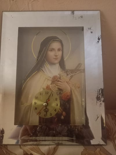 The image shows a framed clock with a religious image of a woman with a halo, holding roses and a crucifix. The clock face is visible, with Arabic numerals and two hands. The frame appears to be made of mirrored glass and is slightly damaged with chipping and smudges. The background is a textured wall in a neutral color. The clock is positioned on a surface that reflects the image, suggesting it might be on a table or shelf. The photograph is taken at an angle, with the clock slightly off-center. There is no indication of the location in Oshane, Bulgaria.