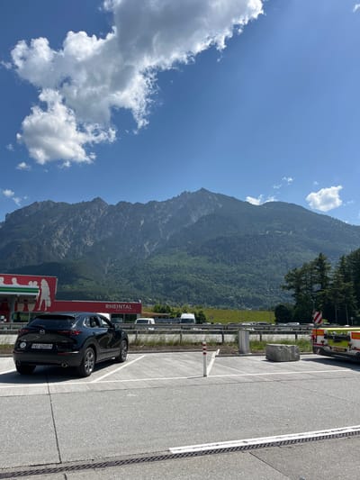 The image depicts a roadside scene in Rans, Switzerland, situated against a backdrop of a large, verdant mountain under a partly cloudy, blue sky. A black Mazda car is parked near a service area marked by a red sign reading "Rheintal Raststätte." The parking area is paved with gray asphalt and marked with white lines. Visible nearby are a guardrail, several vehicles parked in the distance, and trees.