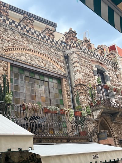 The image shows a section of a stone building facade in Taormina, Italy, with architectural details. The building has a large, multi-paned window with stained glass in shades of green and yellow, and a wrought-iron balcony with several potted plants, including cactuses and hanging greenery. The balcony also features ornate railings. Above the window, the building's facade is decorated with a checkered pattern of black and white tiles and brickwork, resembling a castle battlement. There is another, smaller balcony on the right with similar potted plants and a decorative green ceramic bust. Below the balconies, white awnings are visible, suggesting storefronts or cafes. A sign with Italian text, "VINCENZO DASCANIO," is attached to the building above one of the awnings. The sky is overcast.